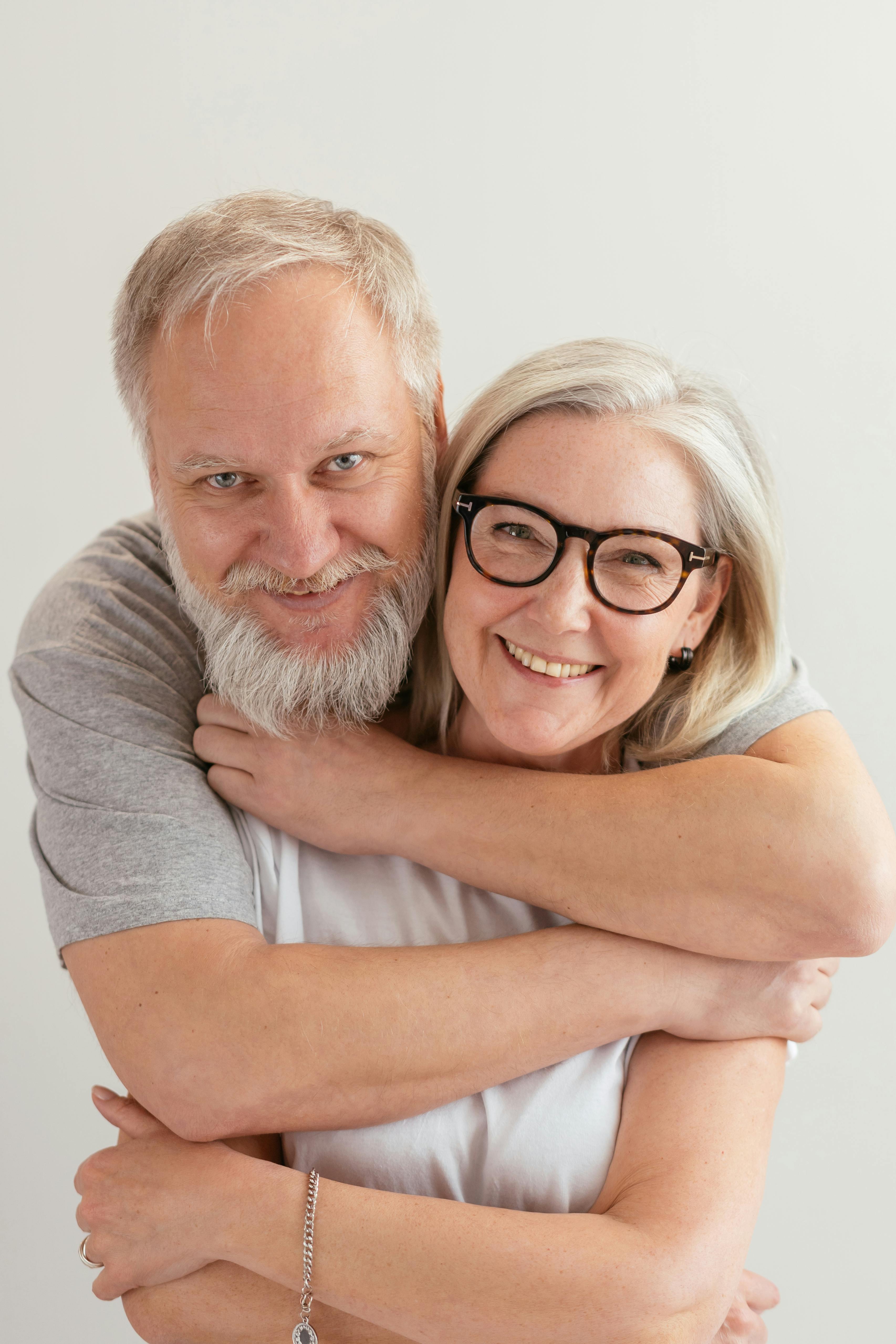 Elderly couple smiling happily at the camera, with the man standing behind his wife, arms wrapped around her, showcasing the joy of a healthy smile after dental implants or dentures from Total Care Dental Boutique in Cairns.